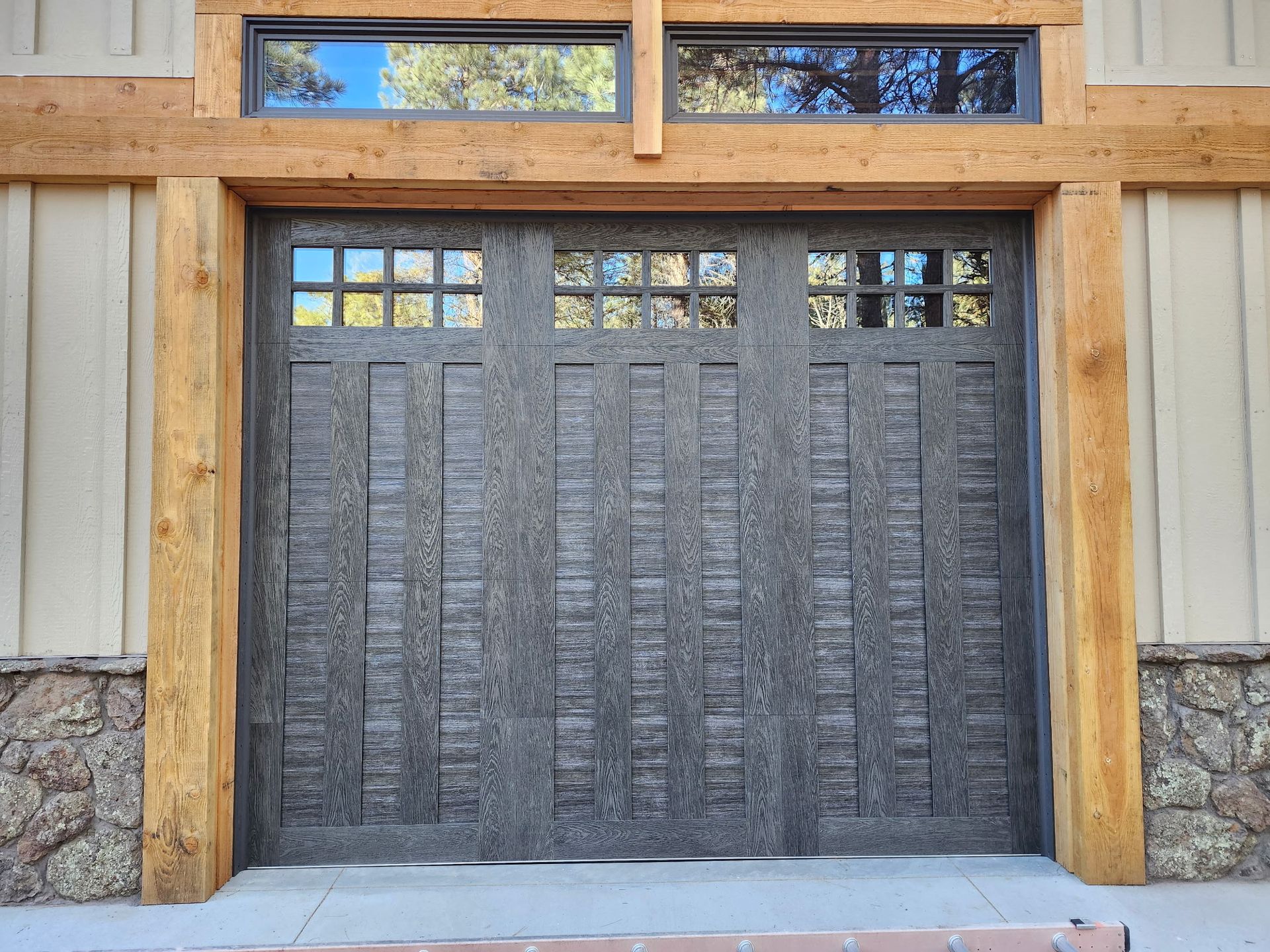 Gray garage door with window panes, framed by wood beams and stone siding.