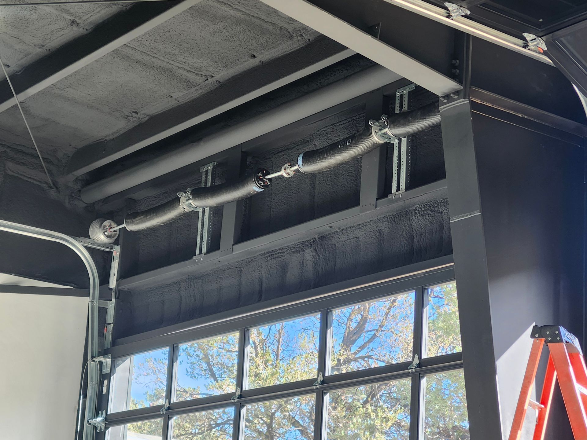 Garage door mechanism with springs and track visible; black frame, light ceiling, and window showing trees.