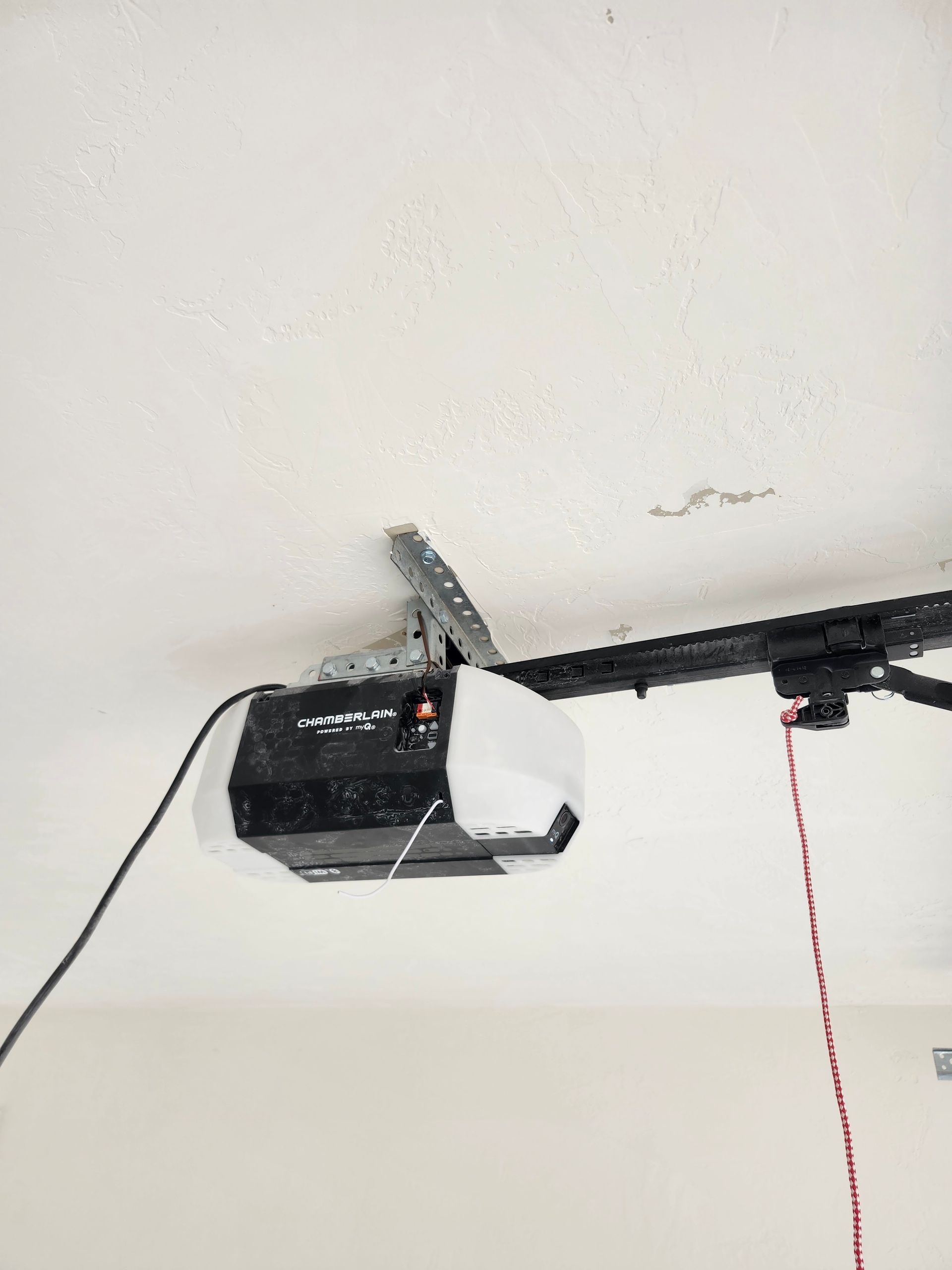 Garage door opener mounted on white ceiling. Black rail extends, red cord hangs.