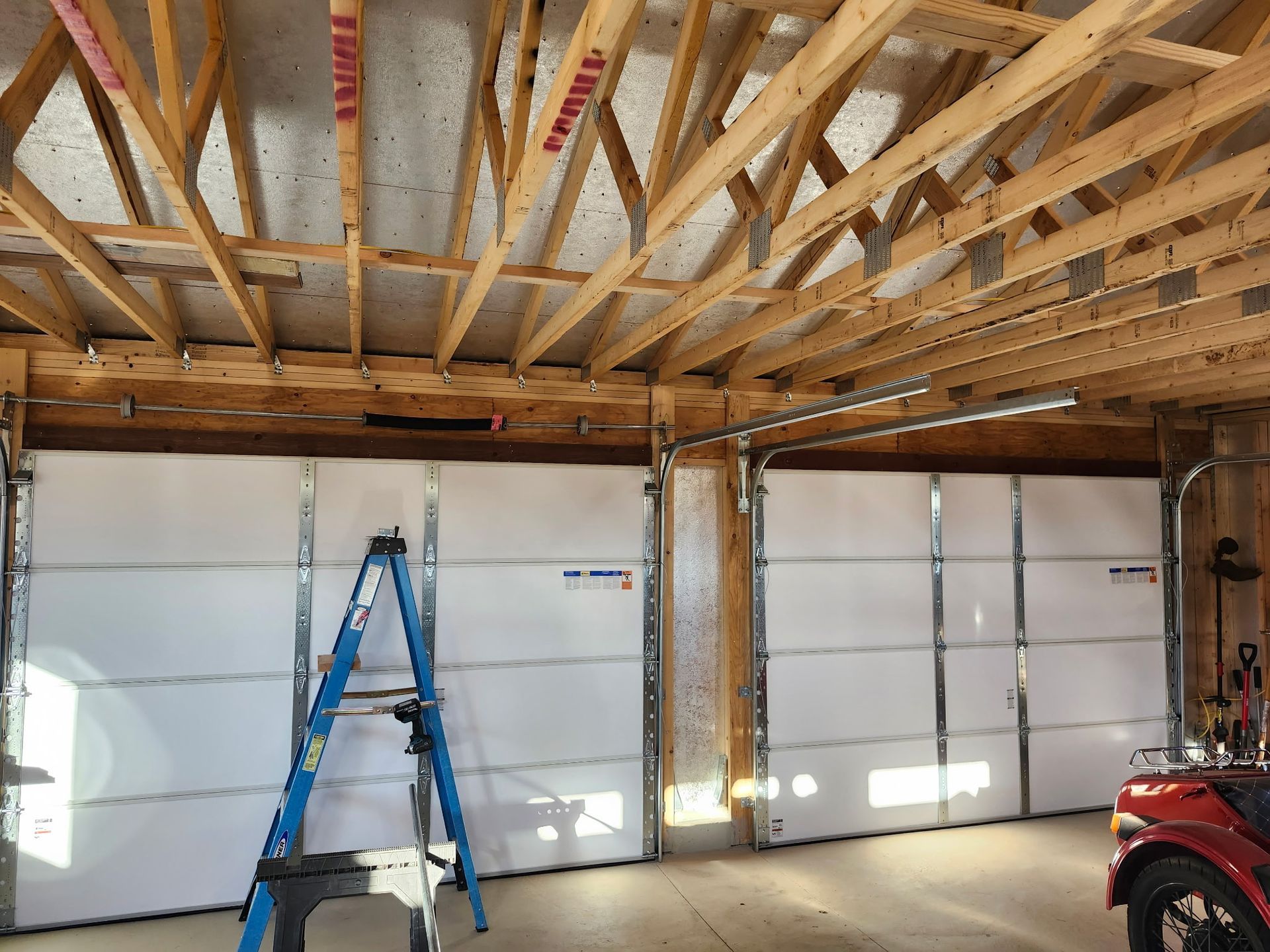 Interior of a garage with two white garage doors and exposed wooden framing. A blue ladder stands nearby.