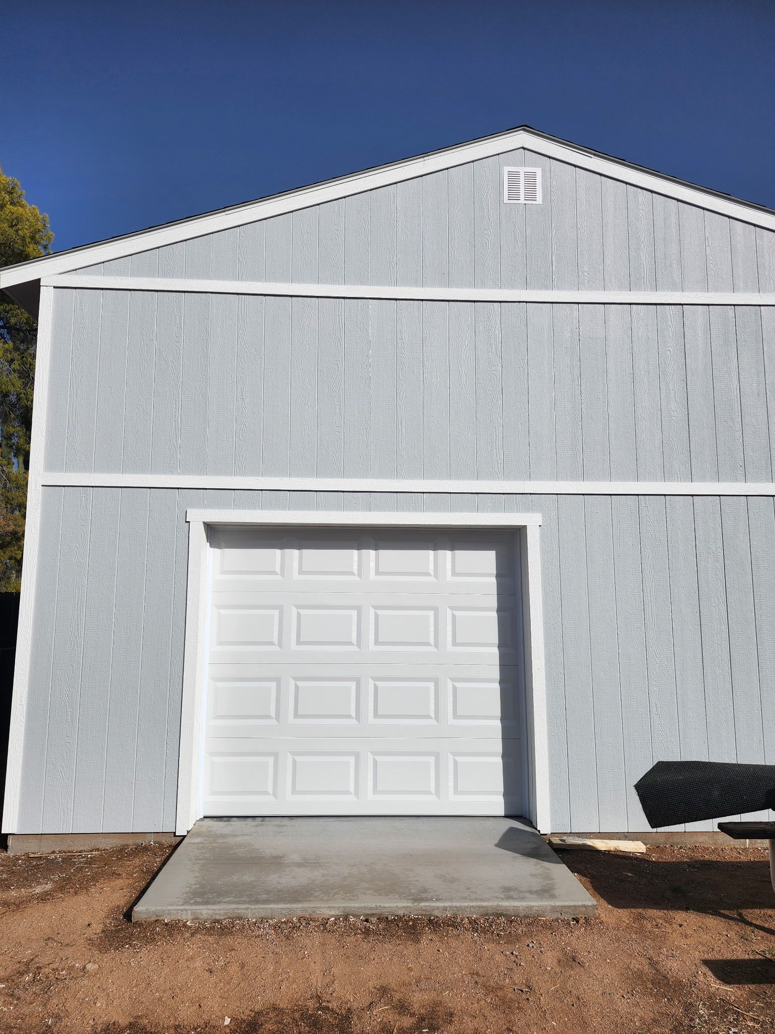 Gray shed with a white garage door, set against a blue sky. Concrete ramp leads to the entrance.