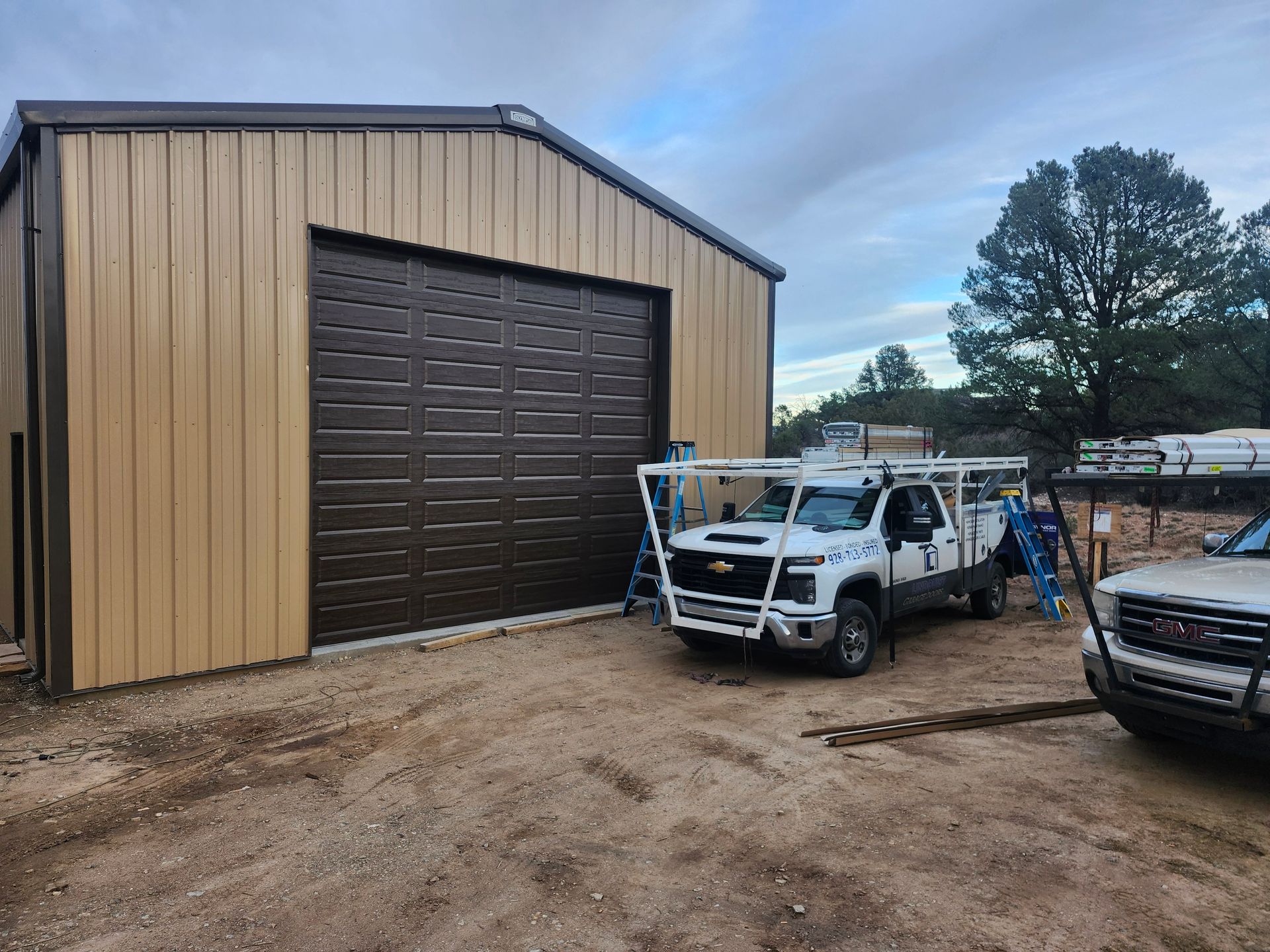 Tan metal building with brown garage door. White trucks parked in front, cloudy sky.