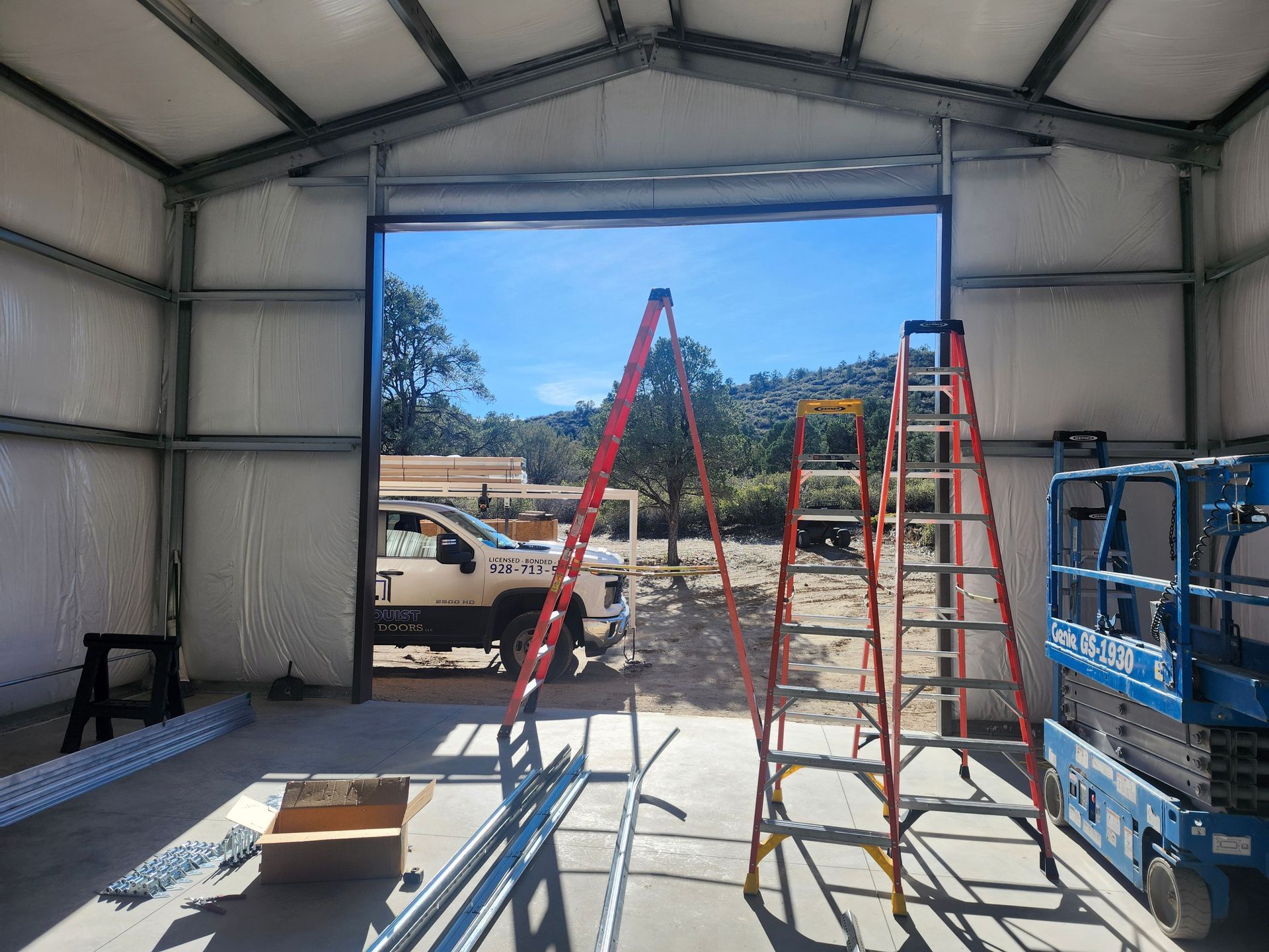 Interior of a metal building under construction, open doorway revealing a sunny outdoor scene, ladders inside.