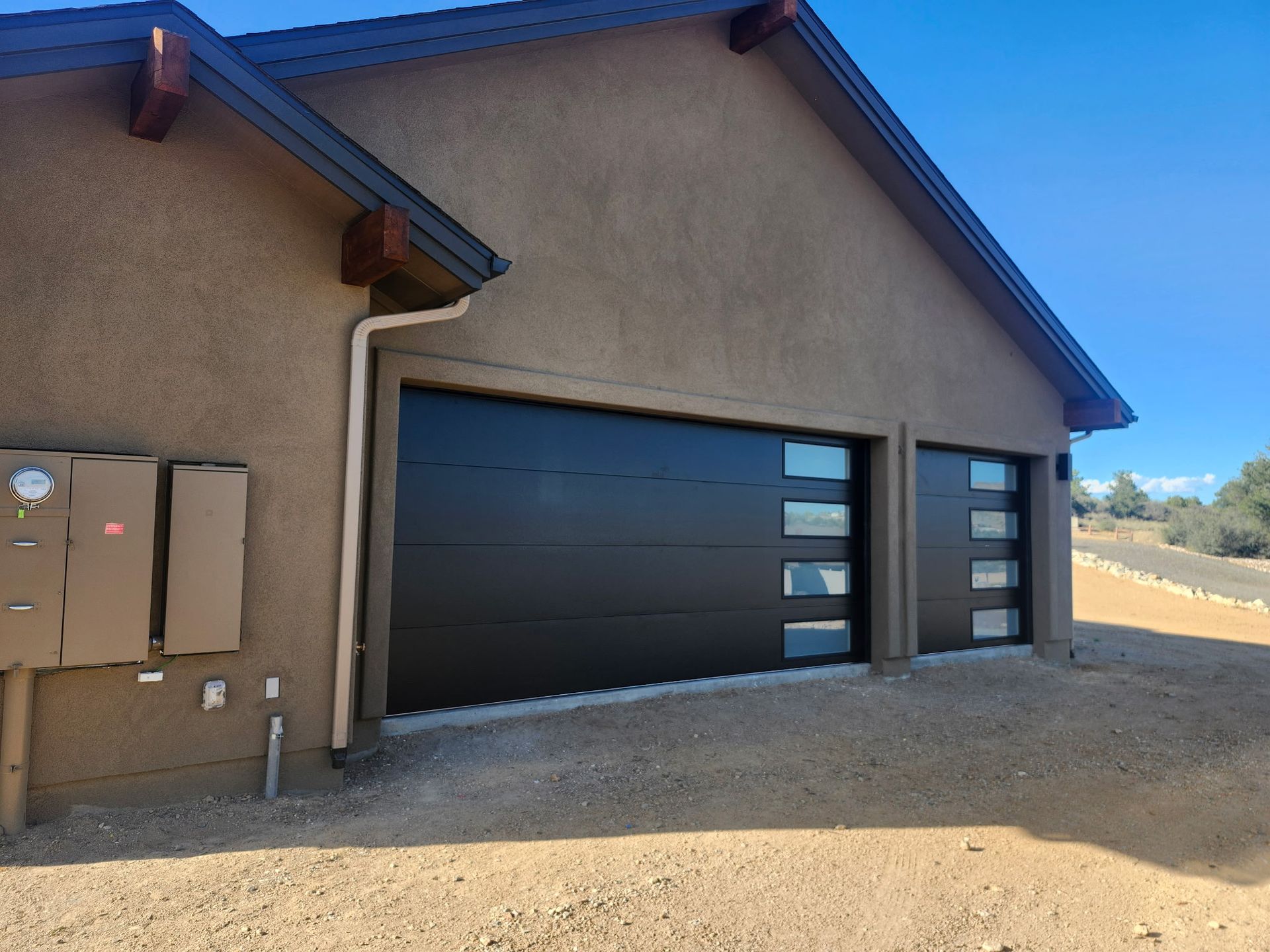 Black garage doors with windows on a stucco building. Brown trim, beige ground, and blue sky.