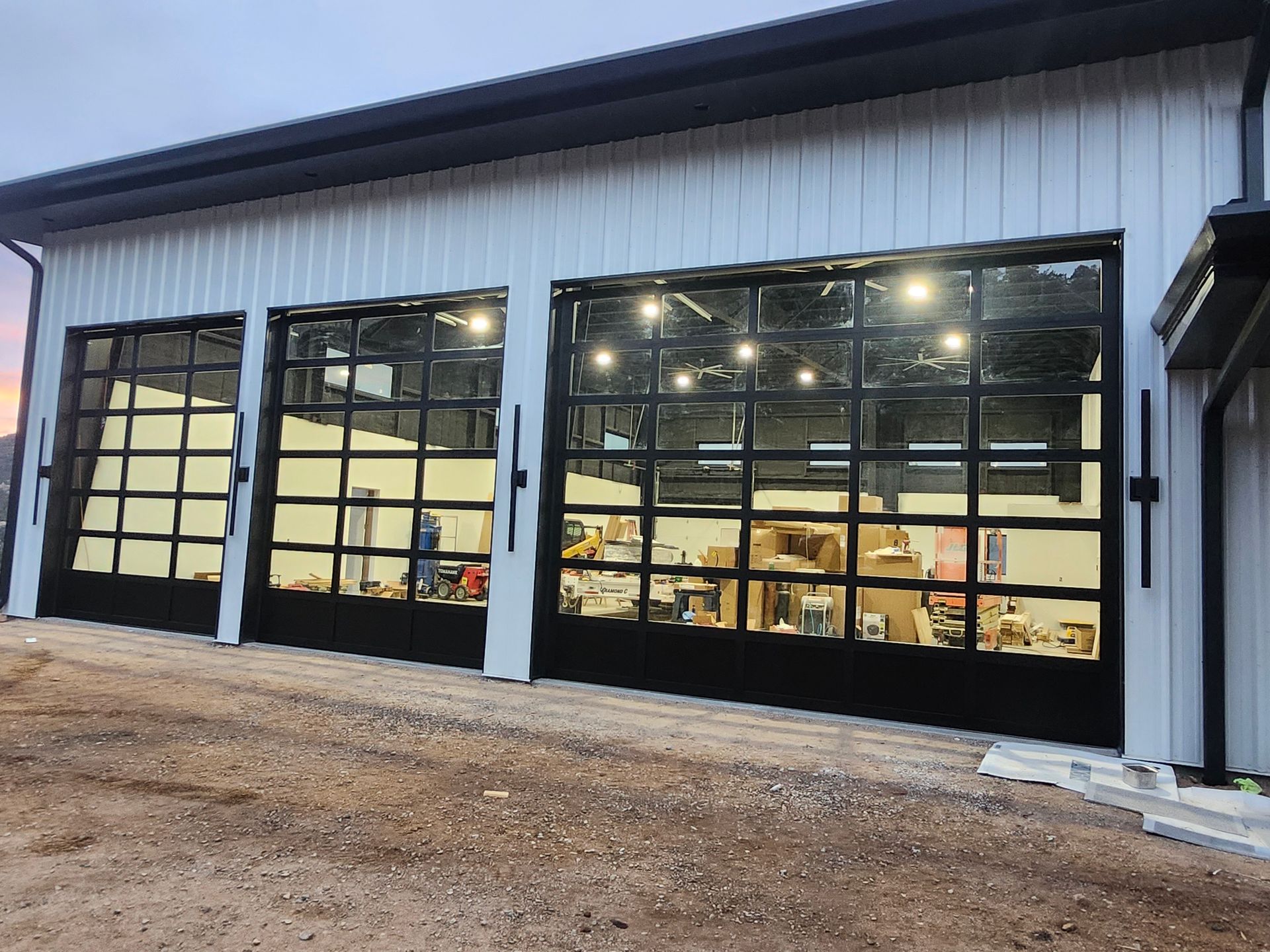 Three large black-framed glass garage doors on a white building, reflecting interior workshop and overhead lights.