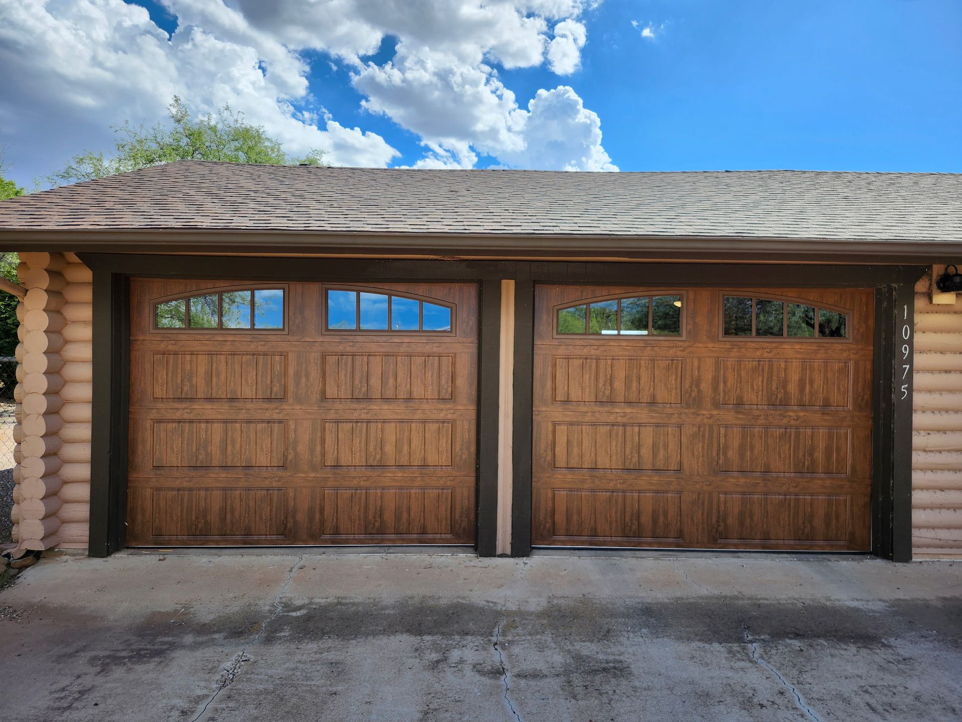 Two brown garage doors with windows, under a wood-shingled roof, against a blue sky with clouds.