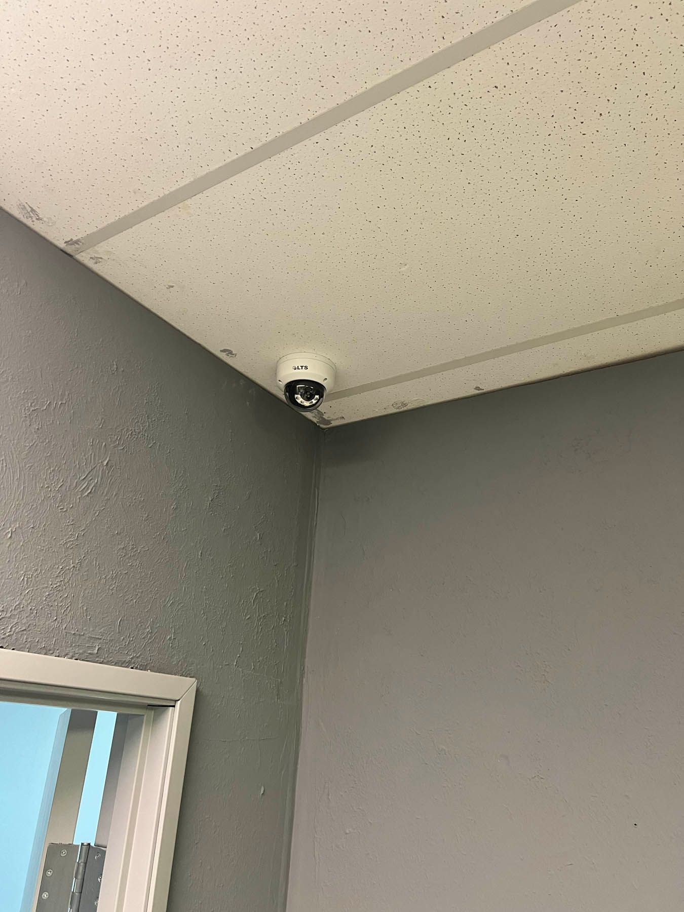 Security camera mounted in the corner of a room, looking down. Gray walls and white ceiling.