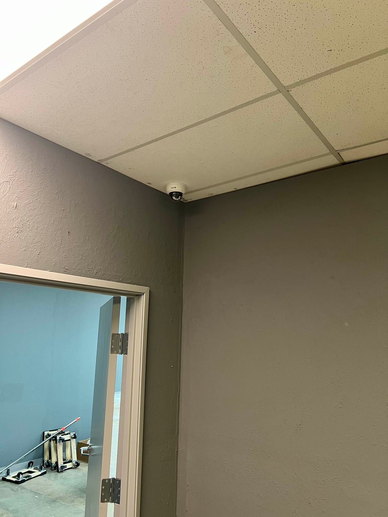 Security camera mounted in the corner of a room, above a door frame. Gray walls and ceiling.