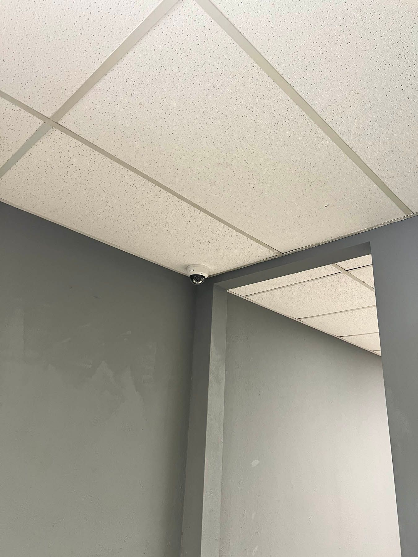 White ceiling tiles and gray walls in a corner, with a security camera mounted in the corner.