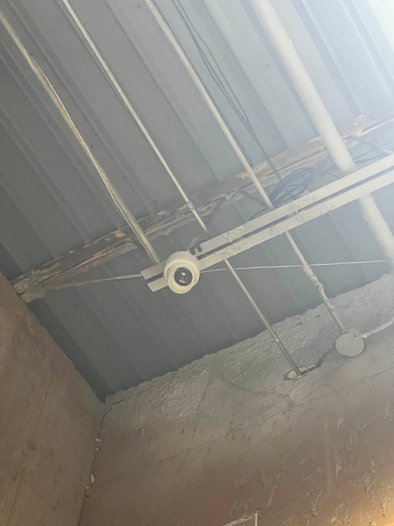 Security camera mounted on a metal ceiling with exposed wires and conduit in a building interior.