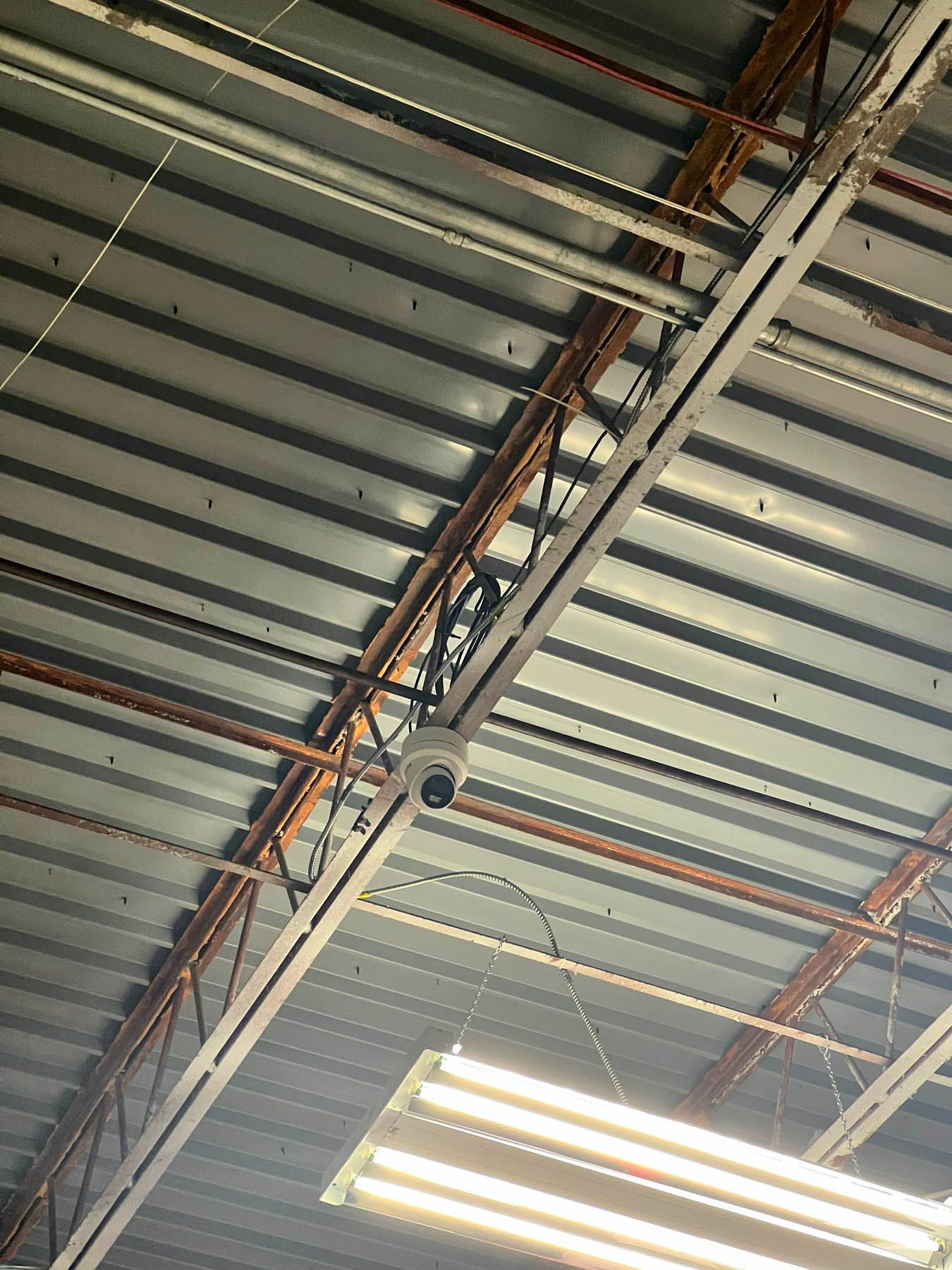 Security camera mounted on a metal ceiling with exposed pipes and lighting.