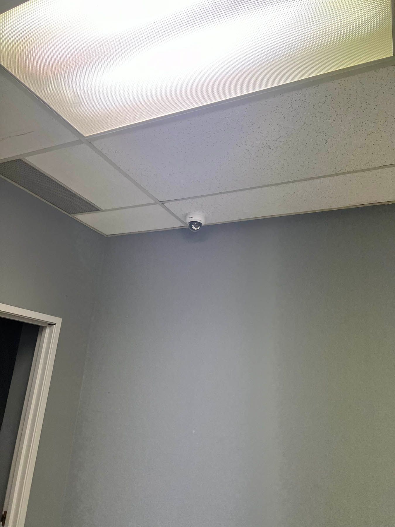 A security camera mounted on a gray wall in a room, with a ceiling light and doorway visible.