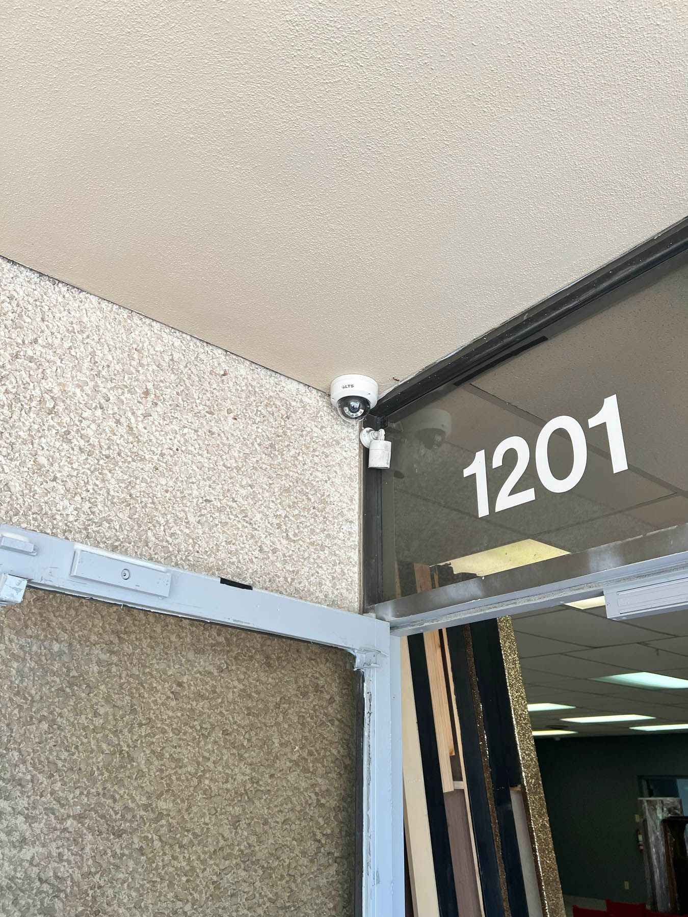 Security cameras outside a building with the number 1201 on the glass door. Beige stucco wall.