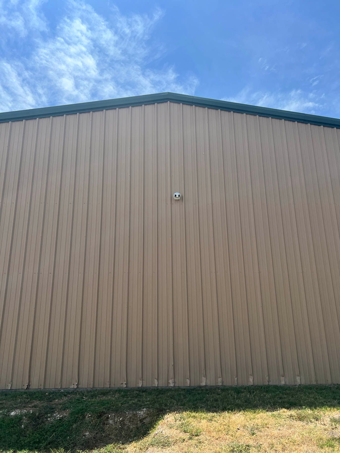 Tan metal building with green trim against a blue sky. A white object is mounted on the wall.