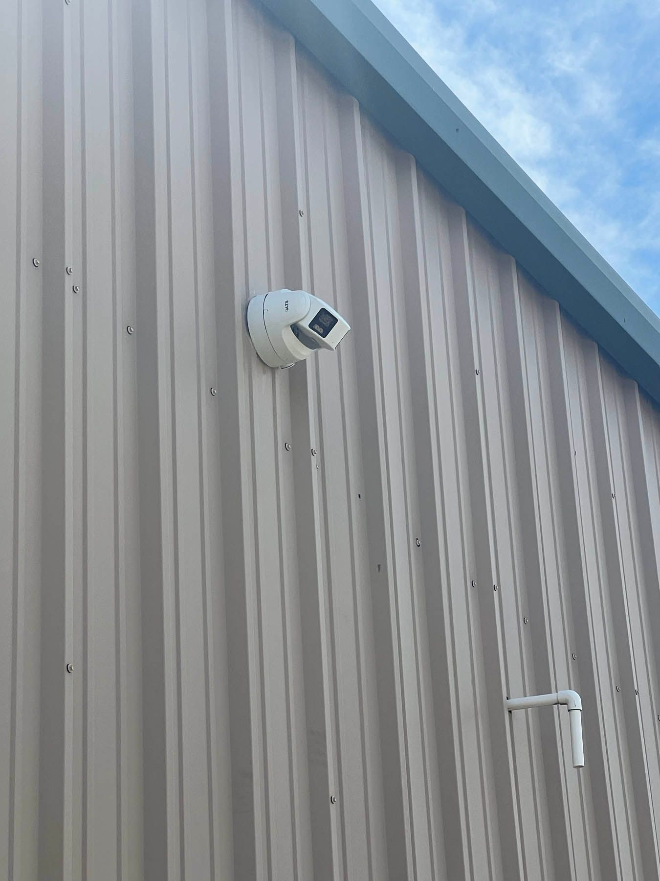 Security camera mounted on a beige corrugated metal building exterior, white housing, and short white pipe.