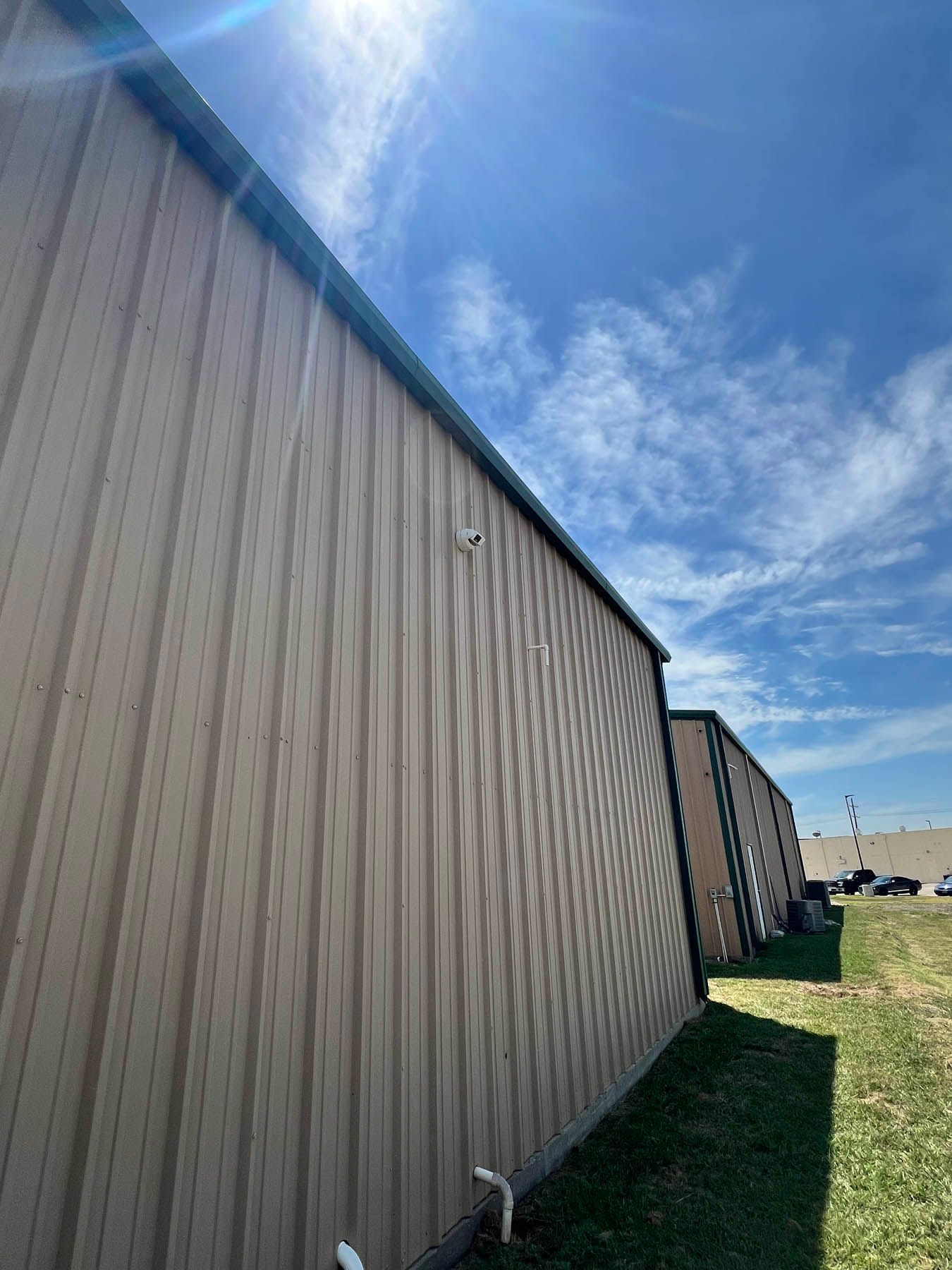 Tan metal-sided industrial building with a blue sky and white clouds. Green grass at the base of the building.