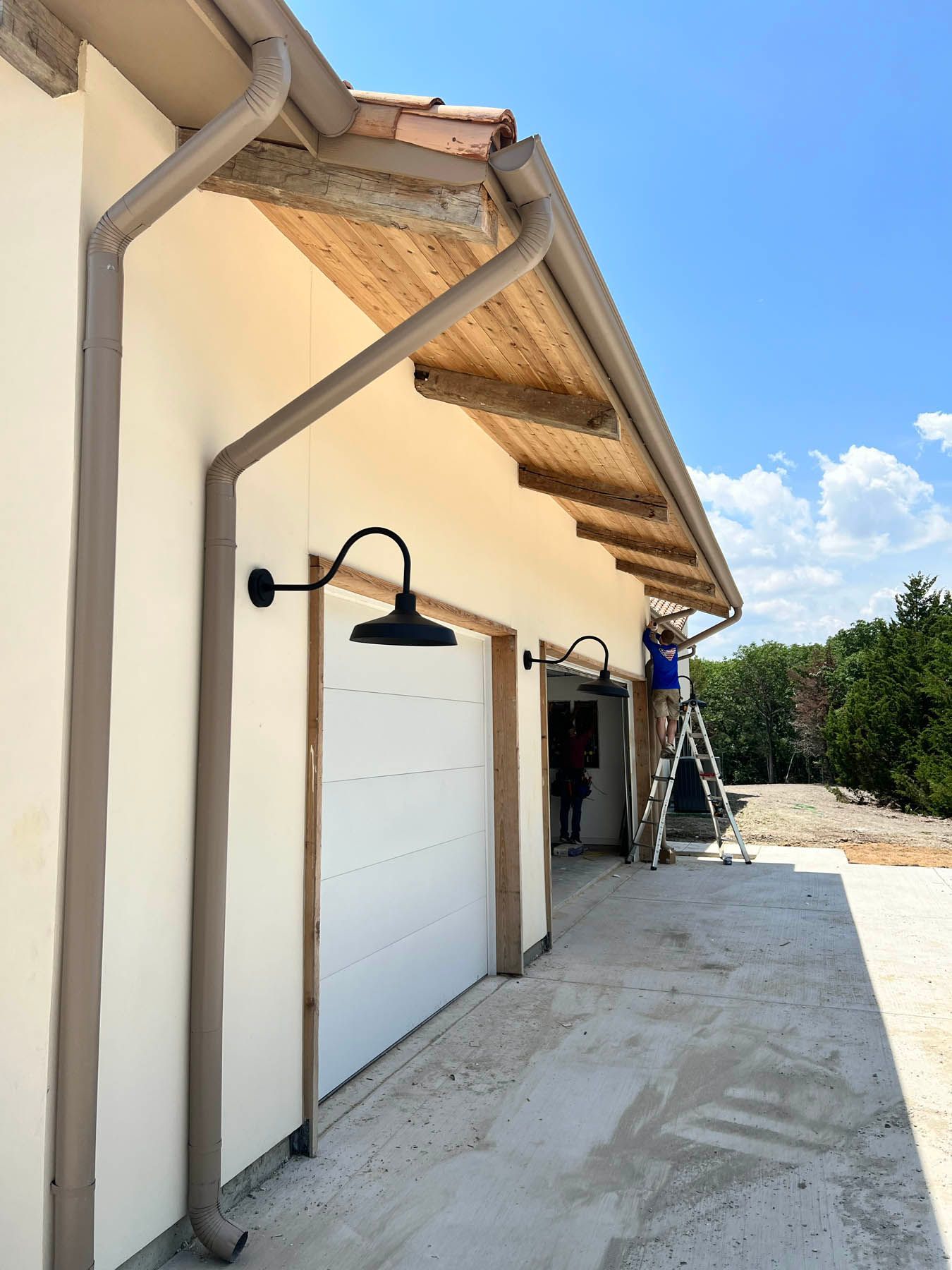 Exterior of a building with a white garage door, black sconce, and a person on a ladder working on the roof.