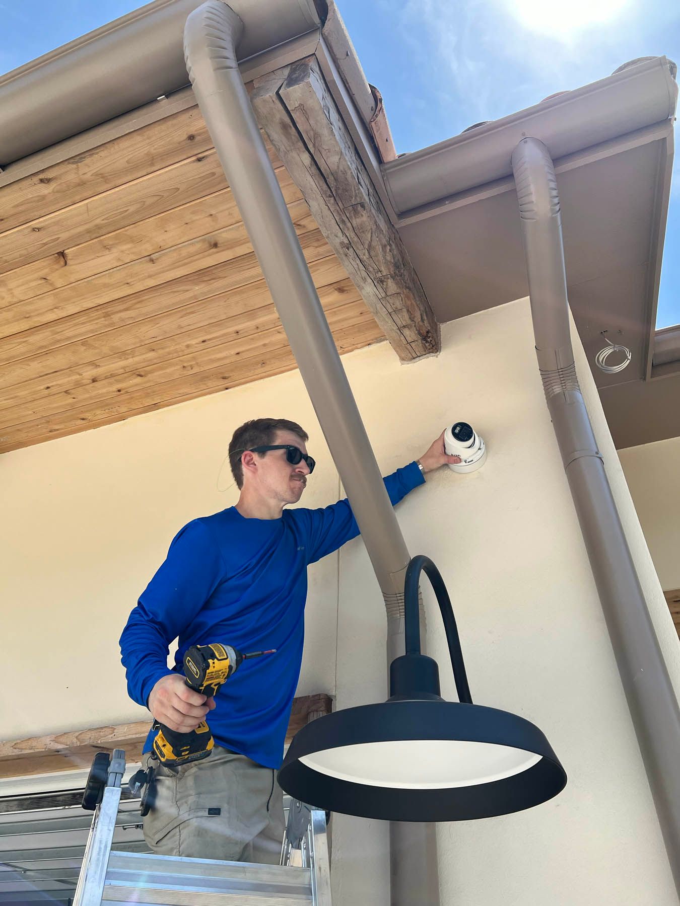 Man on ladder installing a security camera on a building exterior. He holds a drill and wears sunglasses.