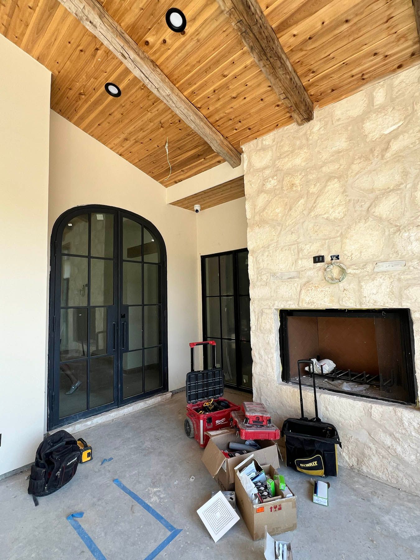 Outdoor patio under construction with stone wall, wooden ceiling, and black-framed doors. Tools are scattered.