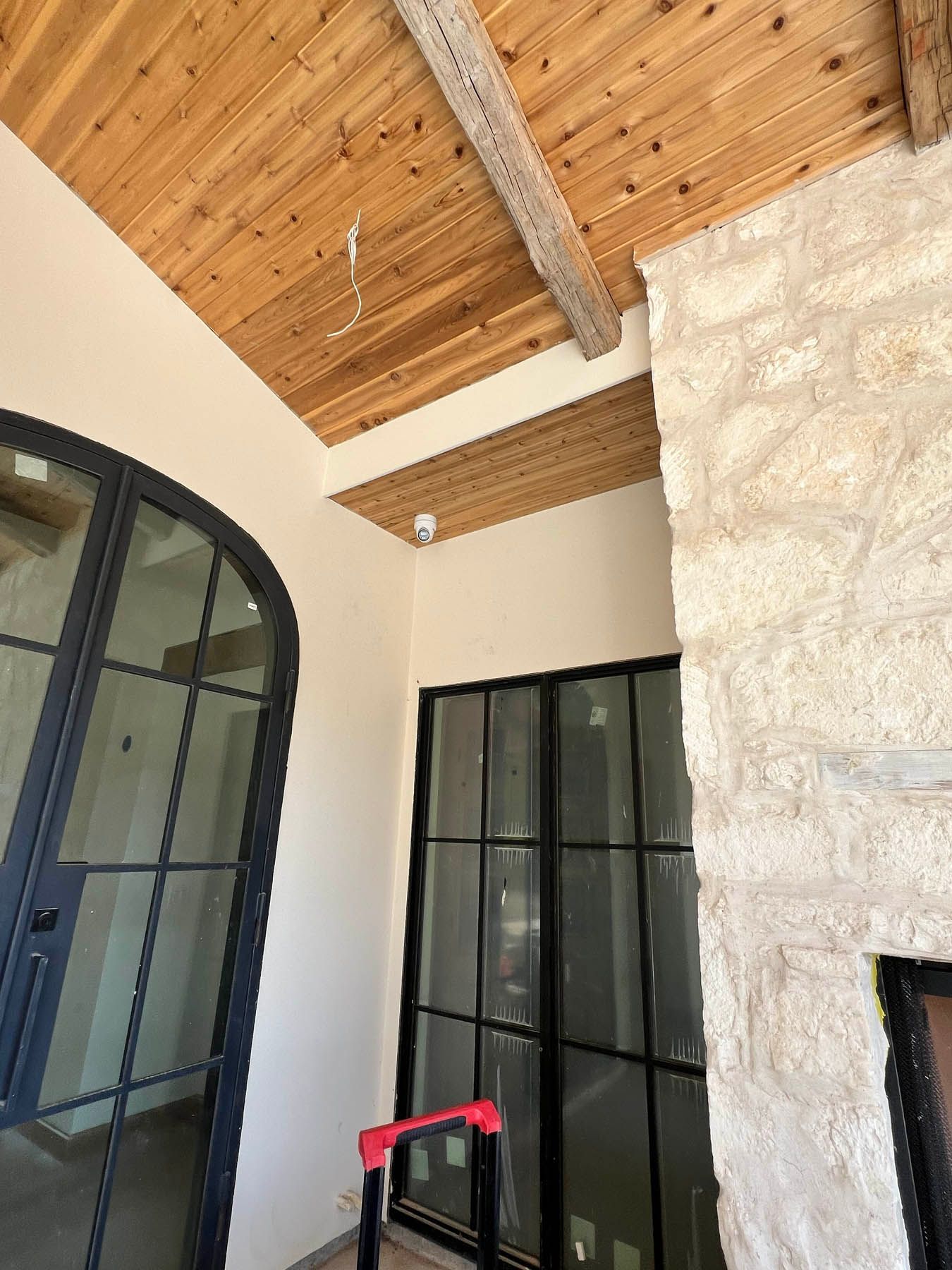 Exterior view of a building with arched black-framed door and wood ceiling. A ladder sits in front of another door.