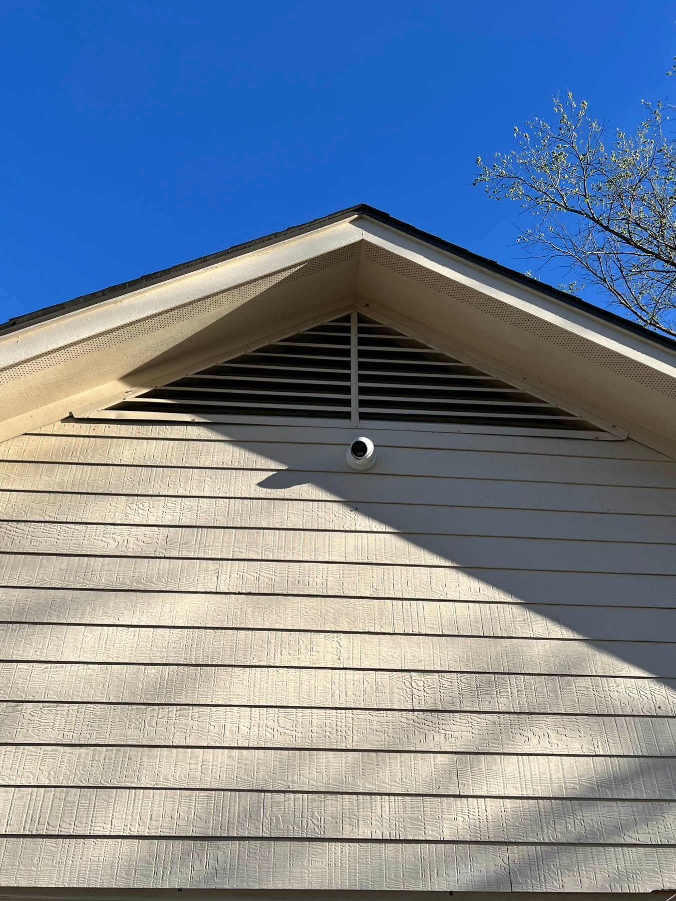 Triangular attic vent with security camera, beige siding, and clear blue sky.