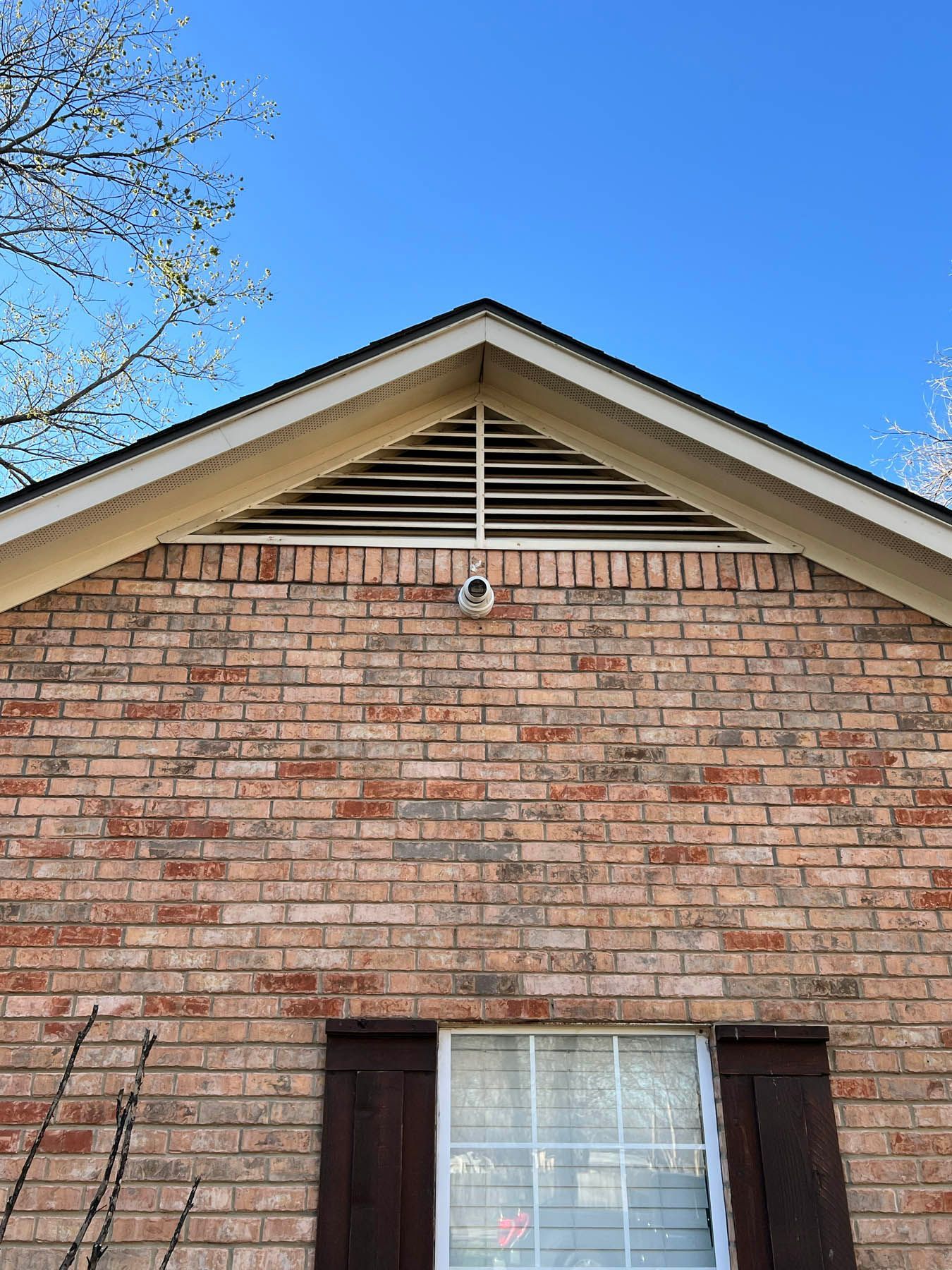 Brick house exterior with a white window and brown shutters. A security camera is mounted under the roof's peak.