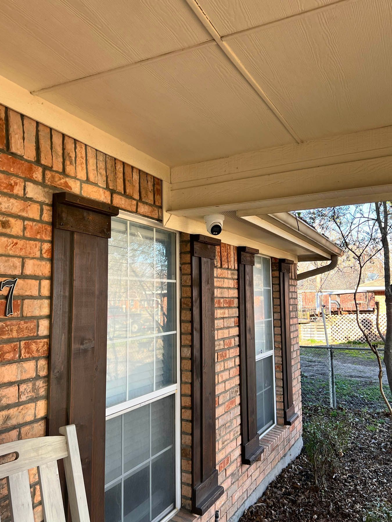 Brick home exterior with windows, dark wood shutters, and a security camera under the porch overhang.