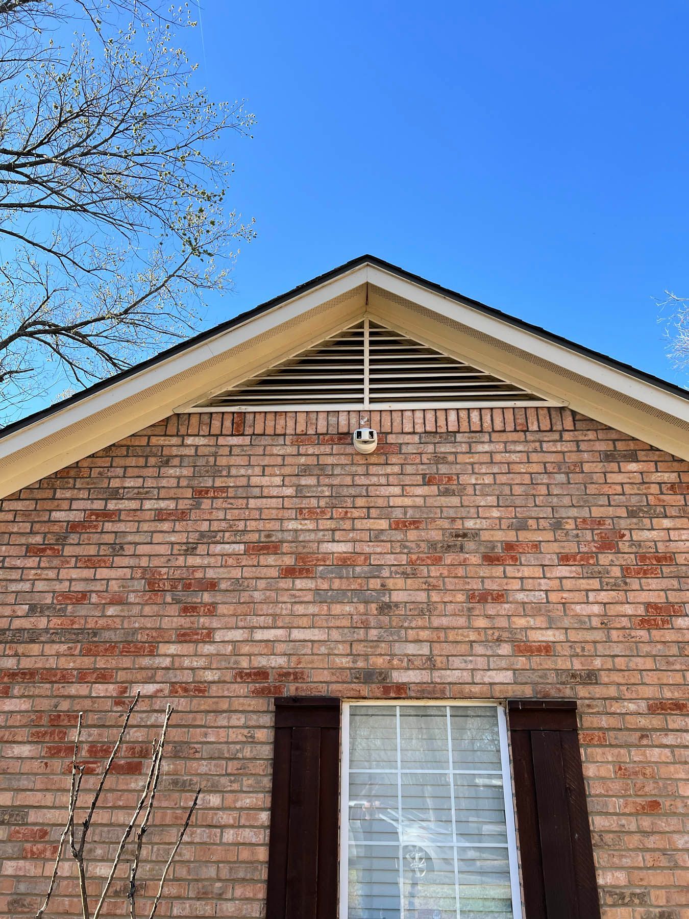 Brick house facade with a peaked roof, vent, and security camera. Window with brown shutters. Blue sky.