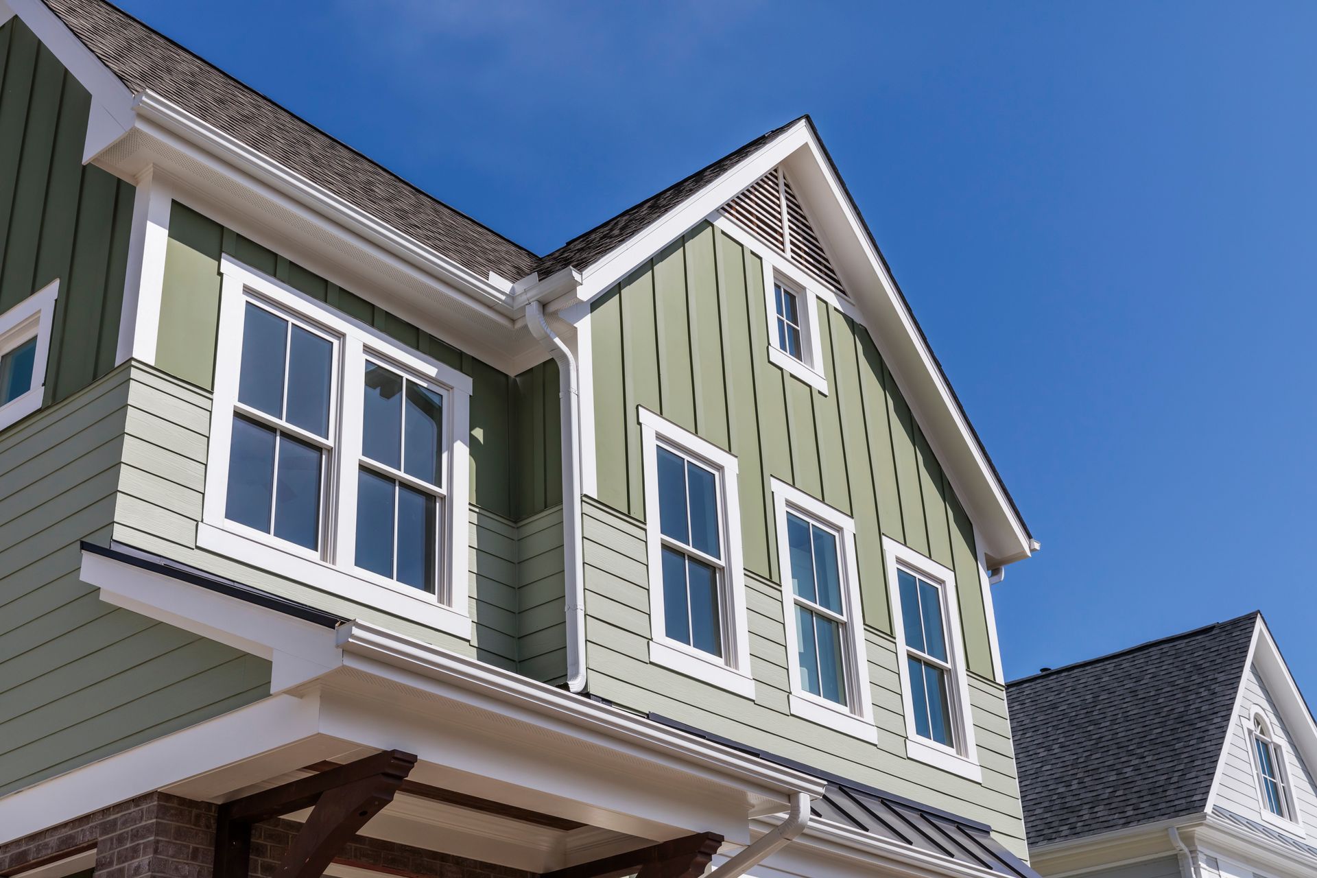 A low-angle view of a sage-green residential house exterior with vertical and horizontal siding and a dark roof.