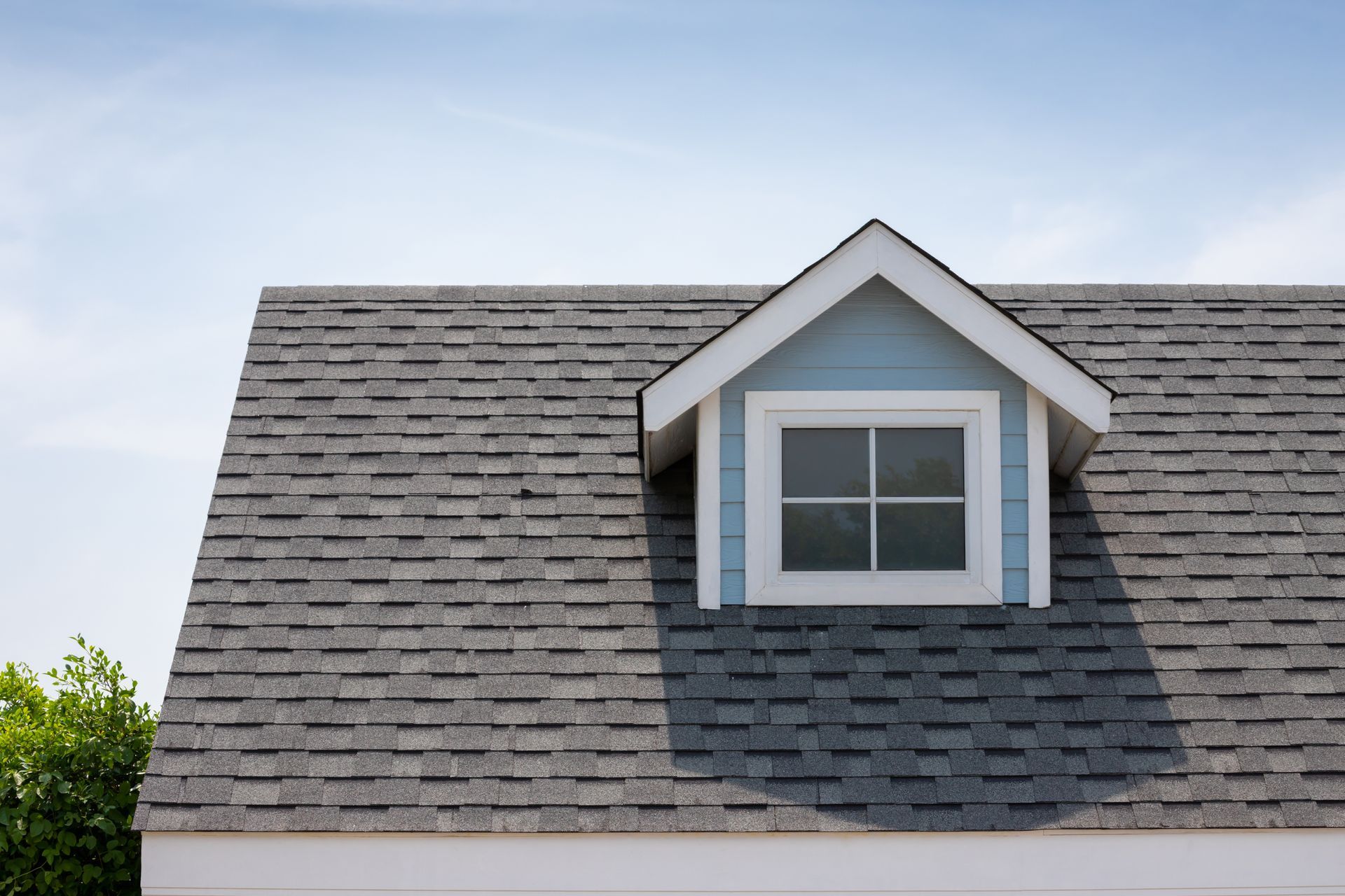A gray shingled roof with a single dormer window featuring light blue siding under a clear blue sky.
