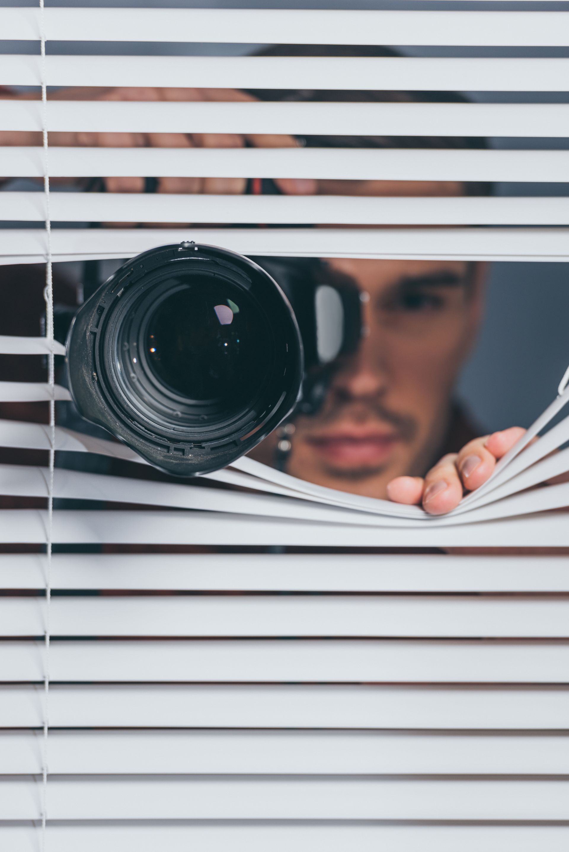 A man is looking through blinds with a camera.