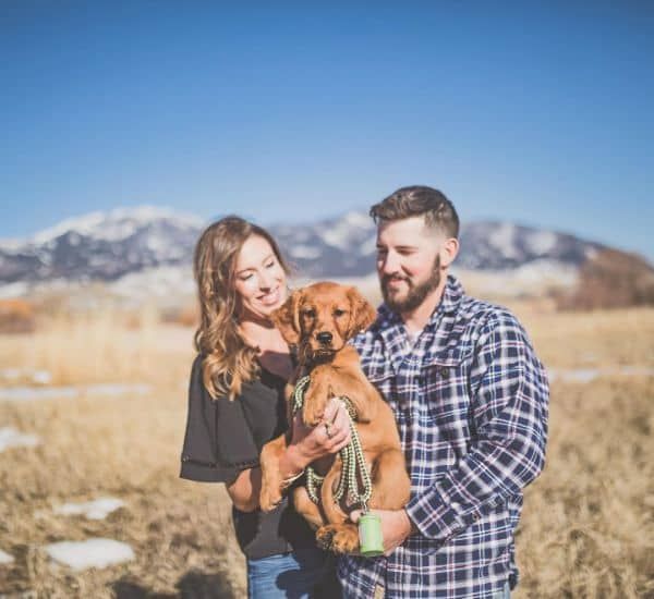Couple holding a puppy in field; mountains in background, sunny day.