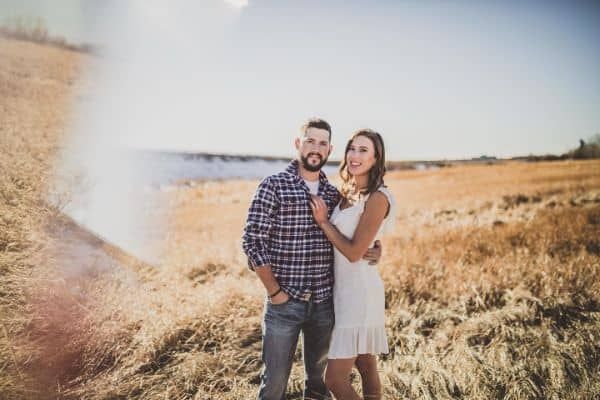 Couple embraces outdoors: man in plaid shirt, woman in white dress. Tall grass field with hazy background.