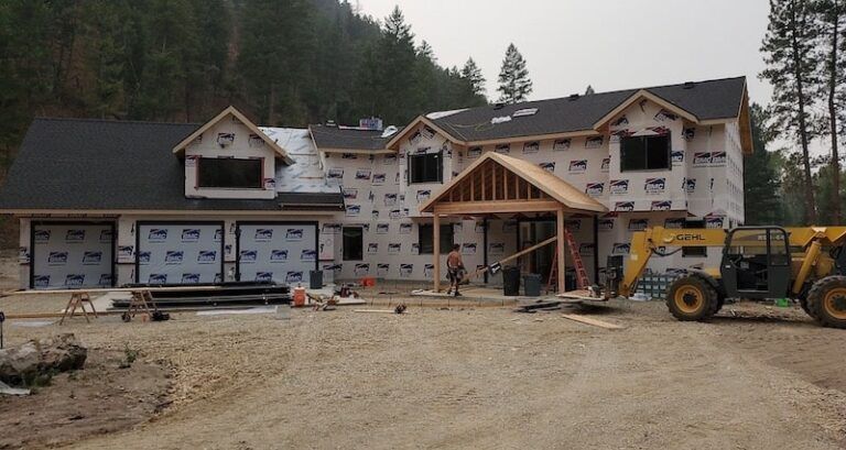 House under construction; covered in paper wrap; garage, entrance, and dormers visible. Dirt lot with heavy machinery.