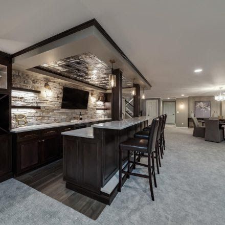 A finished basement with a bar, featuring dark wood cabinetry, stone wall, seating, and a dining area in the background.