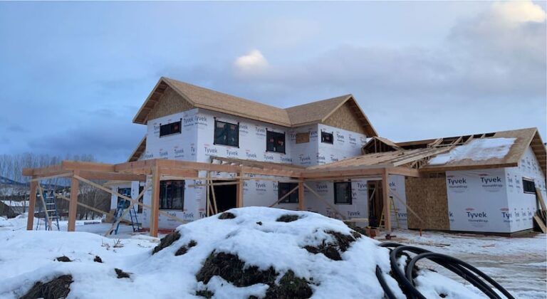 Construction site: two-story house framed with wood, covered in white wrap; snow on ground, cloudy sky.