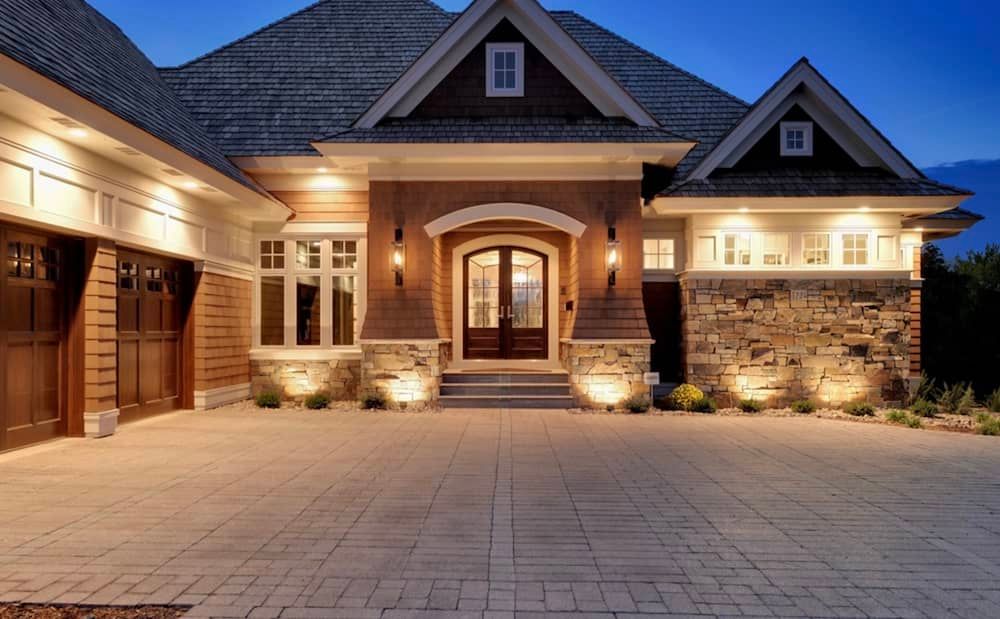 Upscale home exterior with stone, wood, and a brick driveway, illuminated by soft lighting at dusk.