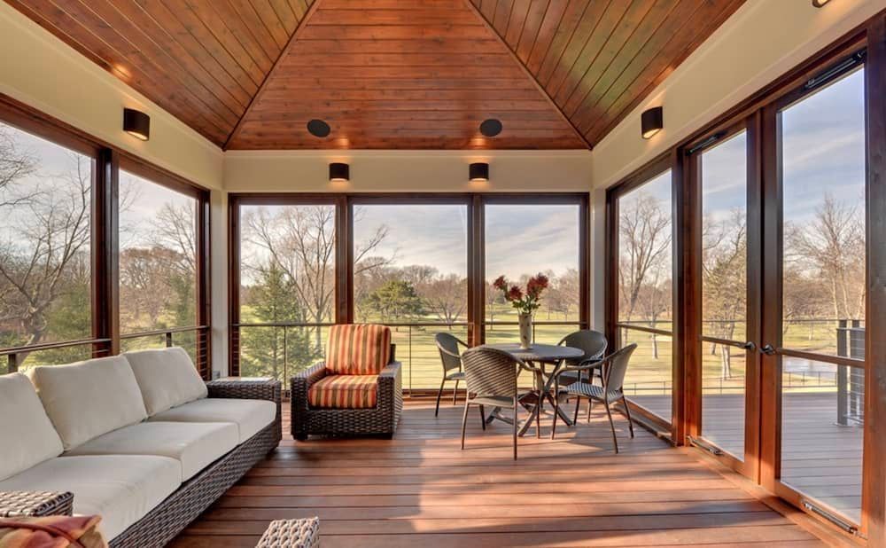 Sunroom with wood floors and ceiling, glass walls, outdoor seating, and a view of trees.