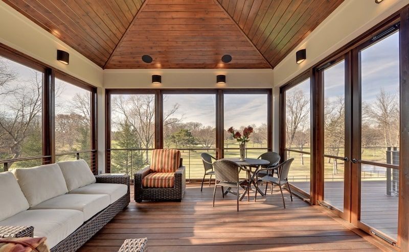 Sunroom with wood ceiling, walls of windows, furniture, and an outdoor view.