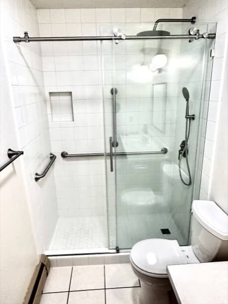 Accessible bathroom with shower, toilet, and grab bars. White tile, glass shower door.
