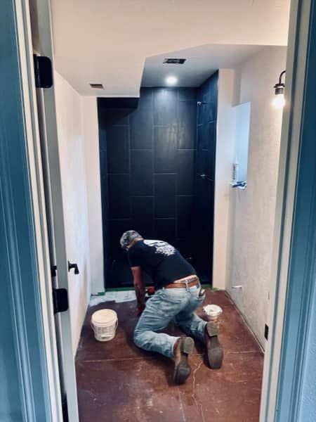 A person kneels on a brown floor inside a bathroom, tiling a black shower wall.