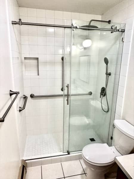 Bathroom with a glass shower, toilet, and white tiled walls. Grab bars for accessibility.