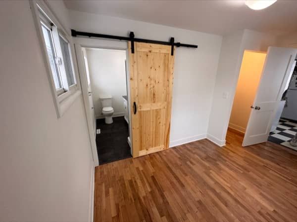 Barn door leads to a bathroom with a toilet. Wooden floors in a room with white walls.