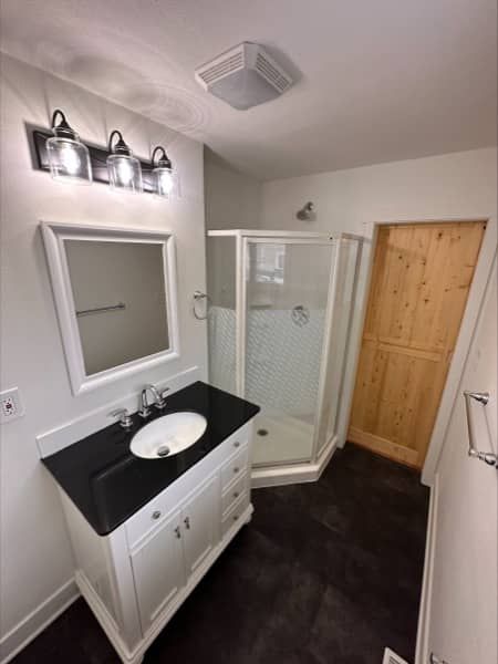 White bathroom with vanity, shower, and light wood door; black countertop, dark floor.