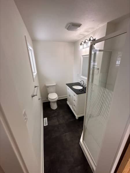 Bathroom with white walls, black floor, toilet, vanity, shower, and a window.
