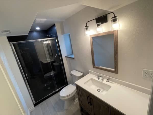 Bathroom with a dark shower, vanity, toilet, and light fixtures. Grey walls and flooring, wood-framed mirror.