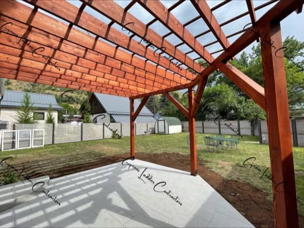 Wooden pergola over a concrete patio, casting shadows on the grass. Houses and trees in the background.