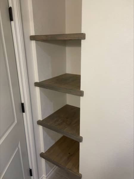 Built-in corner shelving with four wooden shelves; white walls and neutral-colored shelves.