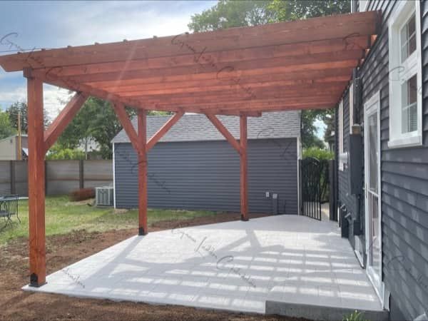 Wooden pergola with a red-brown stain extends over a concrete patio next to a gray house.