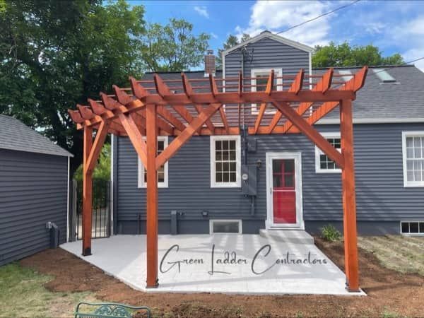 A wooden pergola over a concrete patio in front of a gray house.
